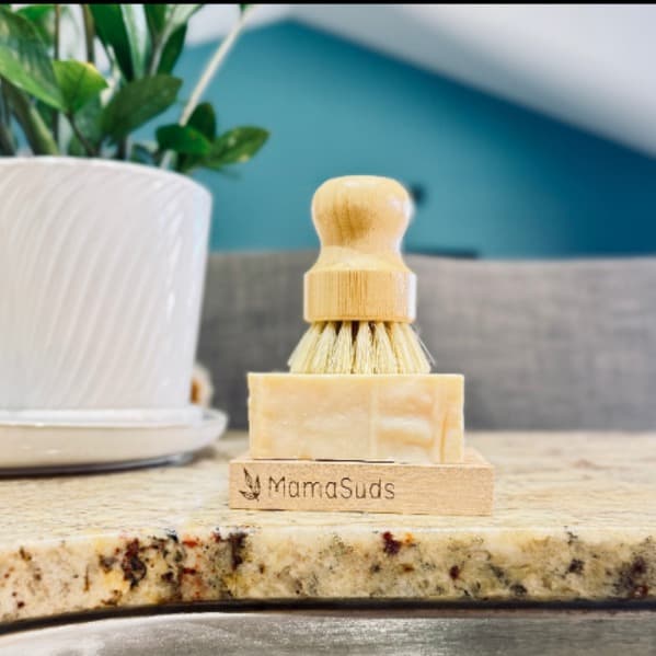 Wooden brush with wooden handle on a stand labeled 'MamaSuds' on a kitchen counter.